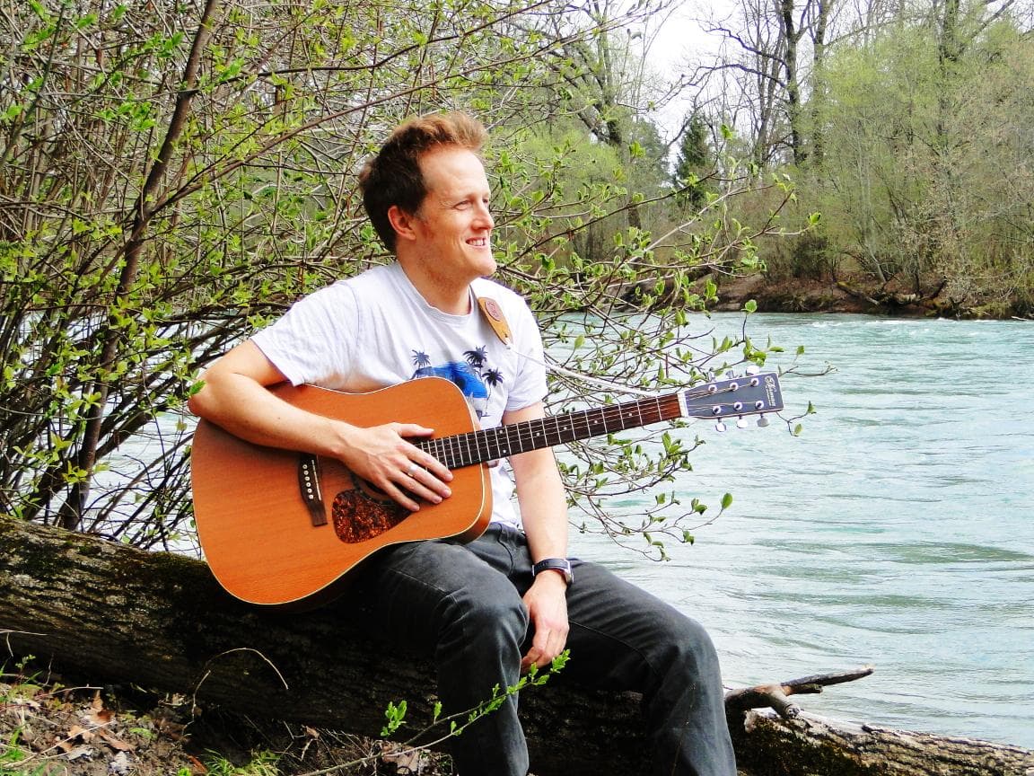 Portrait von Dan Ward, sitzend auf einem Baustamm an einem Flussufer mit einer akkustischen Gitarre umgehängt.